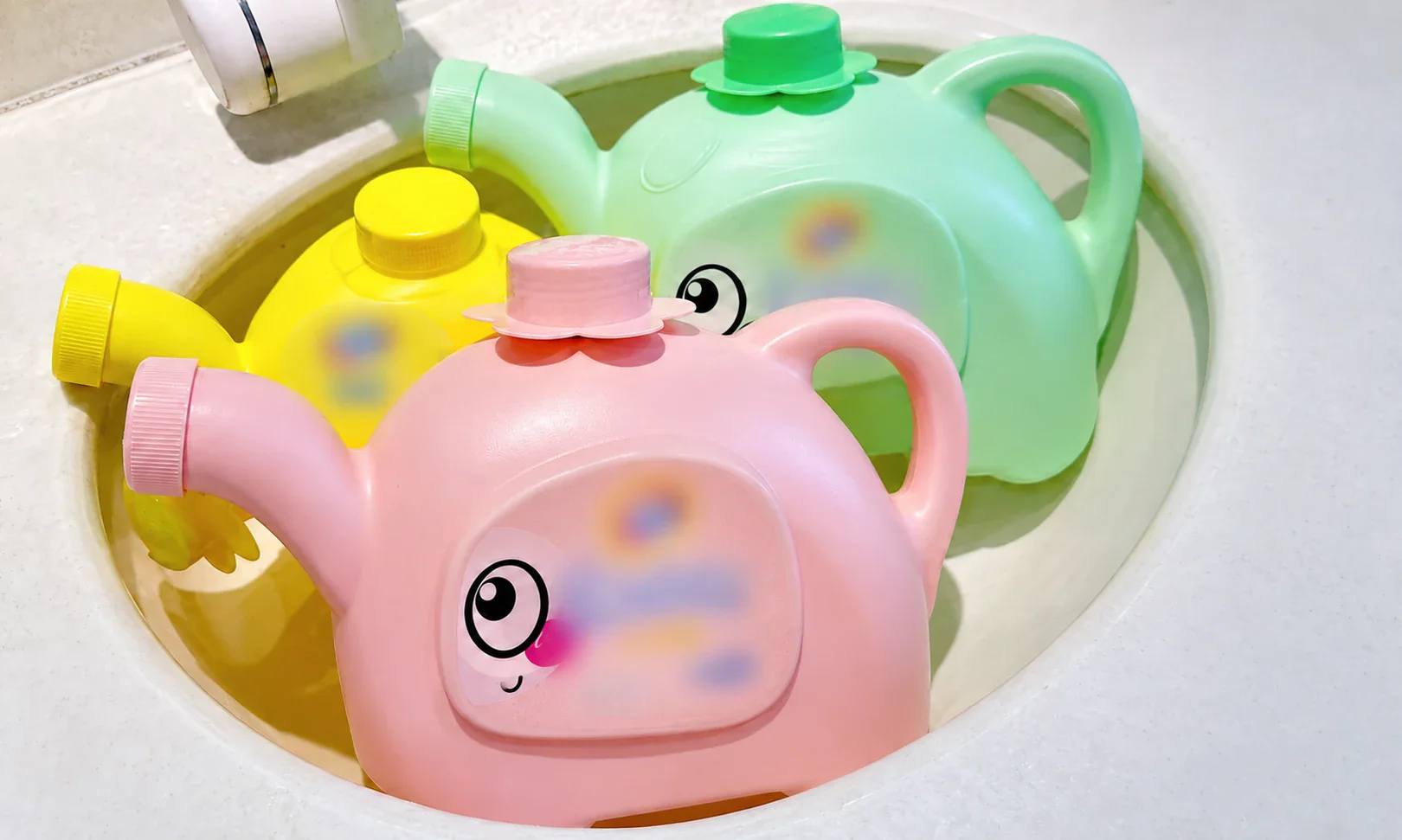 Pink, yellow and green watering-can containers in a sink, with visible handles and spouts and blurred labels